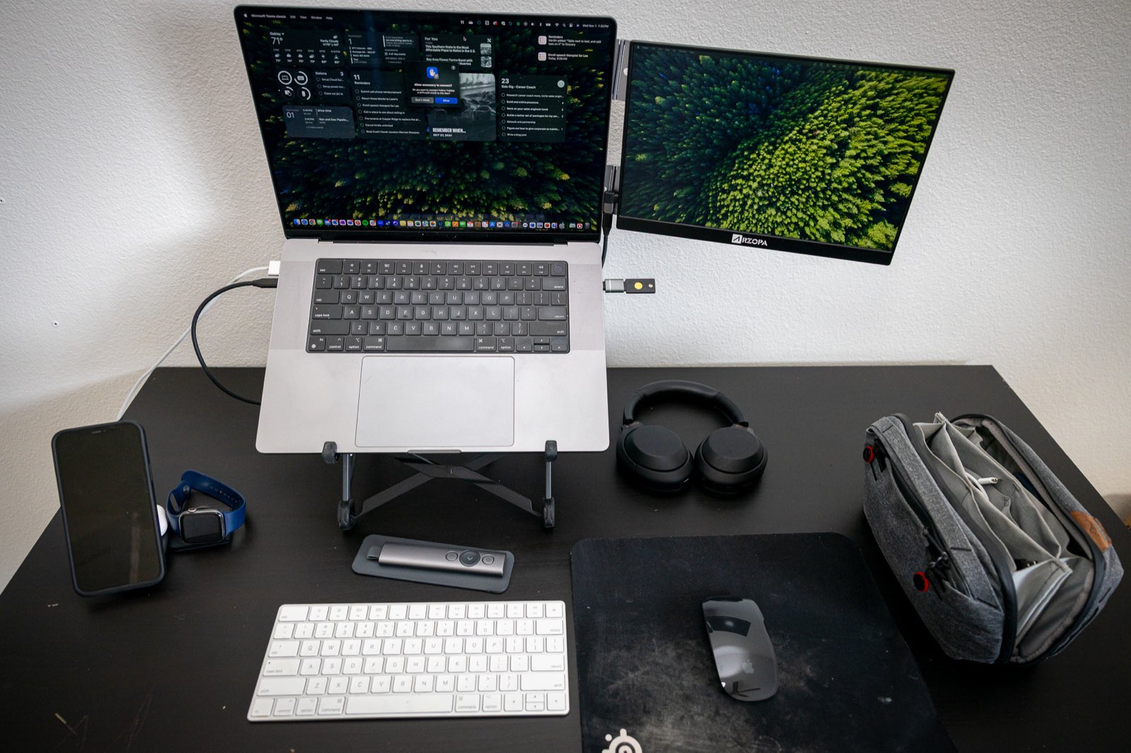 My remote work setup: a MacBook on a Roost stand with a portable second monitor, Magic Keyboard, Magic Mouse, headphones, and a Peak Design pouch on a black desk